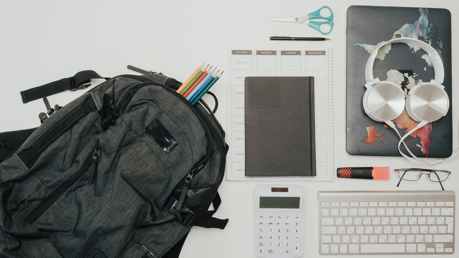 Flat lay of a backpack, notebook, keyboard and headphones on a white desk.